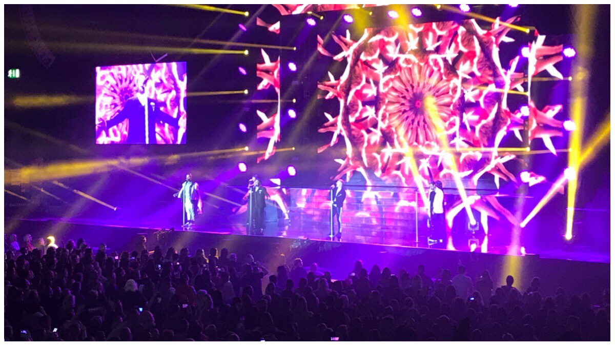 Boyzone on stage at Birmingham arena. The four guys on stage in front of a kaleidoscope of reds and yellow. In the foreground is the crowd in darkness. 